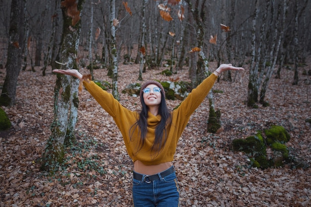 Une femme joyeuse et positive profite de l'automne dans un parc ou dans les bois, porte un pull et un chapeau jaunes, des lunettes. Humeur heureuse hipster girl à la fin de l'automne road trip en forêt