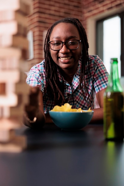 Femme Joyeuse Prenant Des Collations Et Des Boissons Tout En Jouant Au Jeu De Structure De Tour En Bois. Joyeuse Personne Qualifiée Tirant Un Bloc De Bois De La Construction Tout En Profitant D'un Jeu De Société Avec Des Amis à La Maison Dans Le Salon