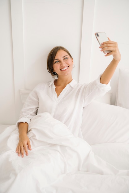 Femme joyeuse prenant un selfie sur un lit blanc