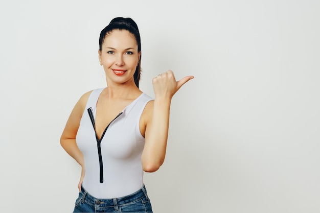 Une femme joyeuse avec une queue de cheval portant un haut blanc sans manches et des jeans denim fait un geste du pouce vers le haut sur un fond blanc qui respire la positivité et l'approbation