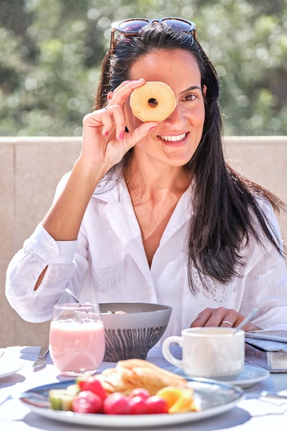 Femme joyeuse regardant à travers un trou dans un donut savoureux tout en se reposant dans la véranda de l'hôtel tout en prenant le petit déjeuner