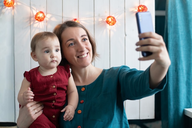 Femme joyeuse et sa petite fille faisant selfie dans la décoration de Noël. Célébration à distance de vacances d'hiver, concept de séjour à la maison. Mère blogueuse créant des histoires.