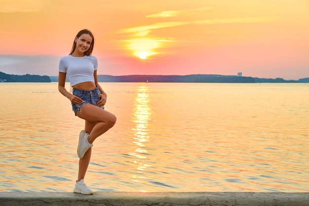 Femme joyeuse en t-shirt et short posant sur une jetée au coucher du soleil