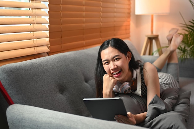 Femme joyeuse tenant une tablette numérique et allongée sur un canapé