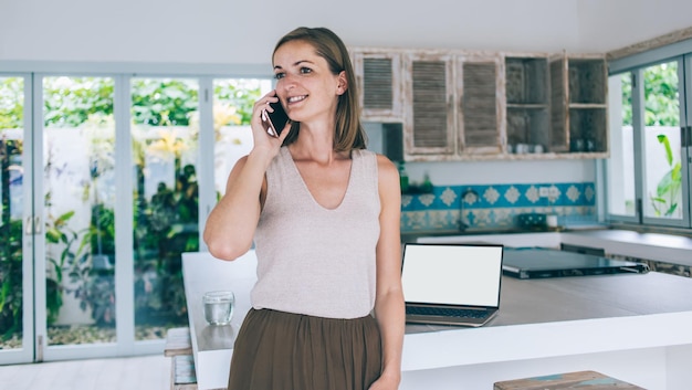 Femme joyeuse en tenue décontractée s'appuyant sur la cuisine ayant un appel sur mobile tout en travaillant sur un ordinateur portable