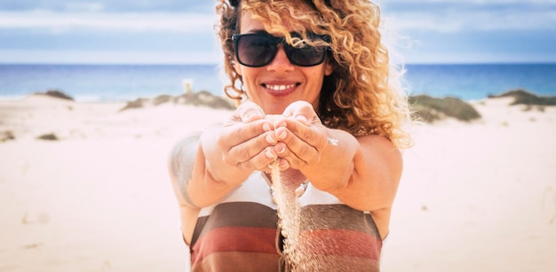 Femme joyeuse en vacances d'été profiter de la plage et du soleil tombant du sable de ses mains et souriant à la caméra Touriste heureux avec l'océan et le ciel en arrière-plan