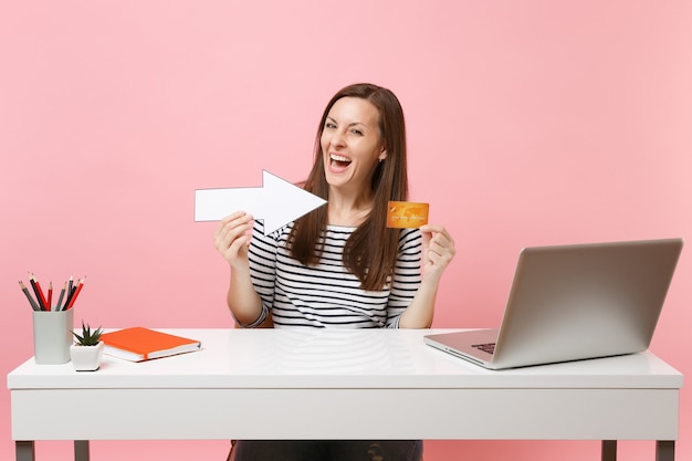 Une femme joyeuse en vêtements décontractés pointant sur une carte de crédit avec une flèche assise travaille au bureau avec un ordinateur portable pc contemporain isolé sur fond rose pastel. Concept de carrière d'entreprise de réalisation. Espace de copie.