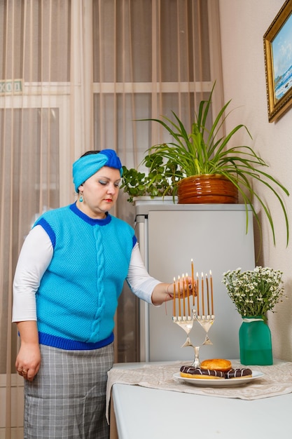 Une femme juive coiffée d'une coiffe dans la cuisine de la salle à manger allume des bougies à Hanoukka sur la table à côté de beignets et d'un vase de fleurs.