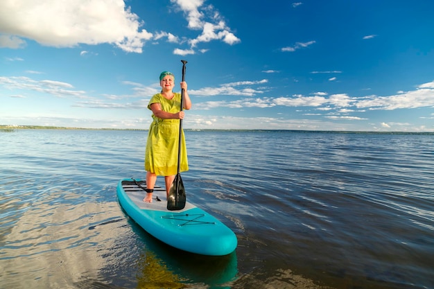 Une femme juive vêtue d'une robe et d'une coiffe debout sur une planche SUP avec une rame flotte sur l'eau contre le ciel bleu