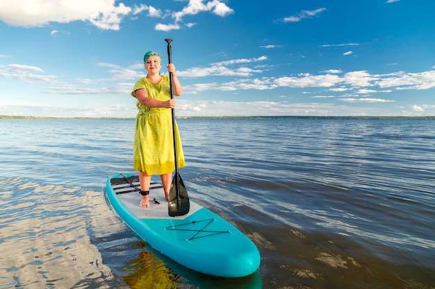 Une femme juive vêtue d'une robe et d'une coiffe debout sur une planche SUP avec une rame flotte sur l'eau contre le ciel bleu