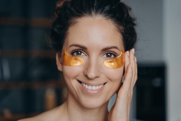 Femme latina souriante appliquant des patchs hydratants dorés sous les yeux à la maison