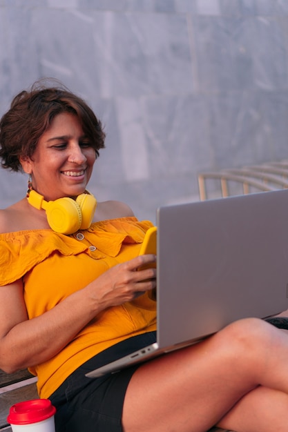 Femme latina utilisant un téléphone portable et un ordinateur portable assis sur un banc dans le parc