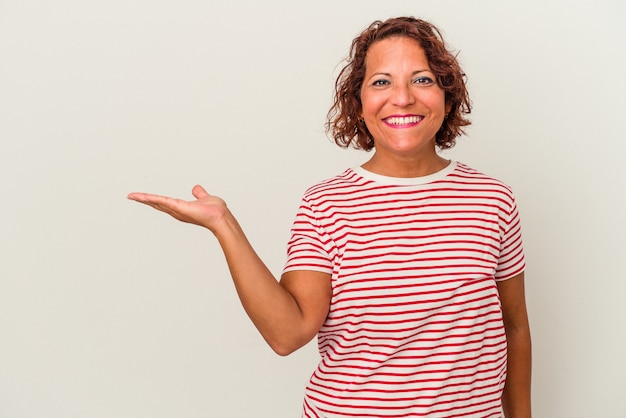 Femme latine d'âge moyen isolée sur fond blanc montrant un espace de copie sur une paume et tenant une autre main sur la taille.