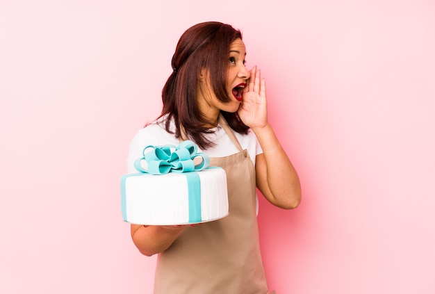 Femme latine d'âge moyen tenant un gâteau isolé sur un mur rose criant et tenant la paume près de la bouche ouverte.