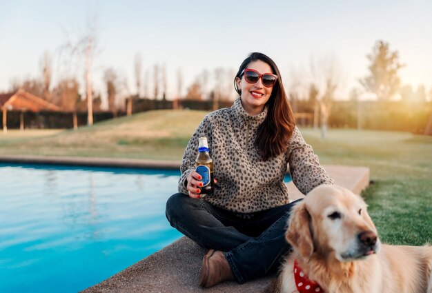 Femme latine assise au bord d'une piscine avec un chien retriever au coucher du soleil