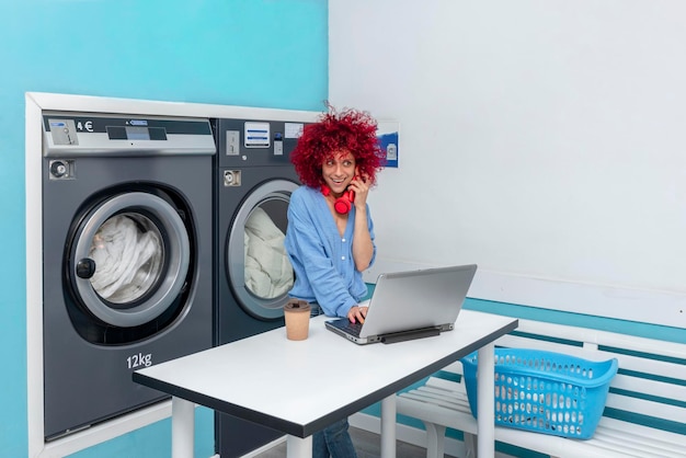 femme latine aux cheveux afro rouges travaille avec un ordinateur portable et parle au téléphone dans la buanderie bleue