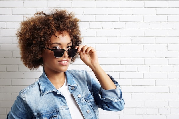 Femme latine aux cheveux bouclés portant des lunettes de soleil et une veste en jean sur un mur de briques