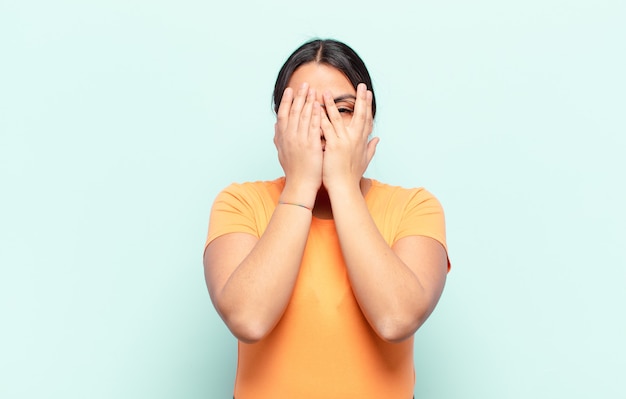 Femme latine couvrant le visage avec les mains, regardant entre les doigts avec une expression surprise et regardant sur le côté