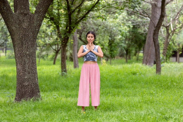 Femme latine faisant la méditation sur le côté d'un arbre dans le parc sur l'herbe verte avec une tenue rose