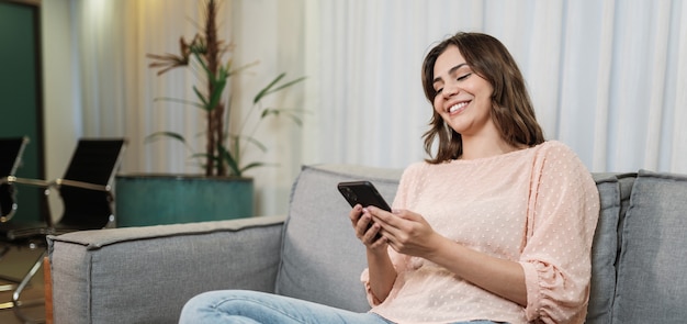 Une femme latine heureuse se détend seule à la maison, assise dans la chambre, partage de bonnes nouvelles sur les réseaux sociaux via son téléphone portable. Une femme souriante profite du week-end pour commander des aliments en ligne dans l'application téléphonique.