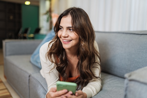 Une femme latine heureuse se détend seule à la maison dans la chambre et partage de bonnes nouvelles sur les réseaux sociaux via son téléphone portable. Une femme souriante profite du week-end pour commander des aliments en ligne dans l'application téléphonique.