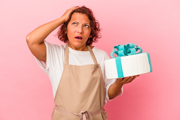 Femme latine de pâtisserie d'âge moyen tenant un gâteau isolé sur fond rose étant choquée, elle s'est souvenue d'une réunion importante.