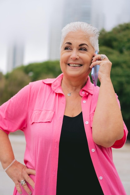 Photo femme latine plus âgée aux cheveux blancs lors d'un appel téléphonique souriant dans un parc vêtue de rose