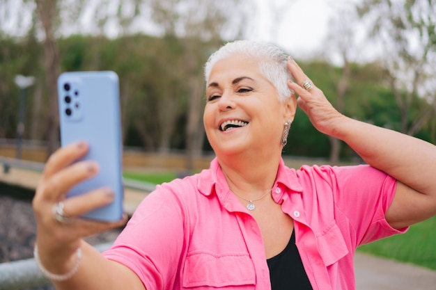 Photo femme latine plus âgée aux cheveux blancs prenant un selfie souriant avec un téléphone portable dans un parc vêtu de rose