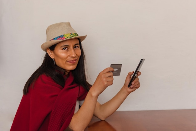 Femme latine portant un chapeau et un châle rouge assis tenant son téléphone portable et une carte de crédit sur fond blanc