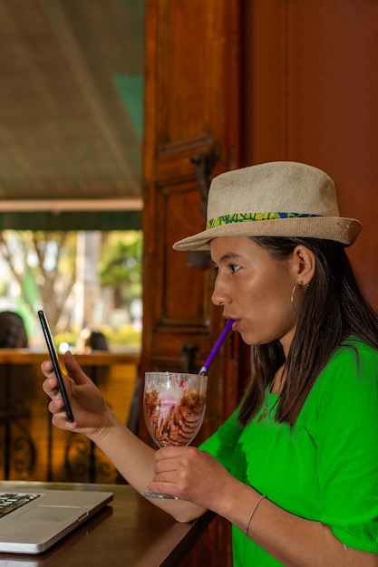 Femme latine portant un chapeau et un chemisier vert assis dans un vieux café vérifiant son téléphone portable tout en buvant un milk-shake avec une paille