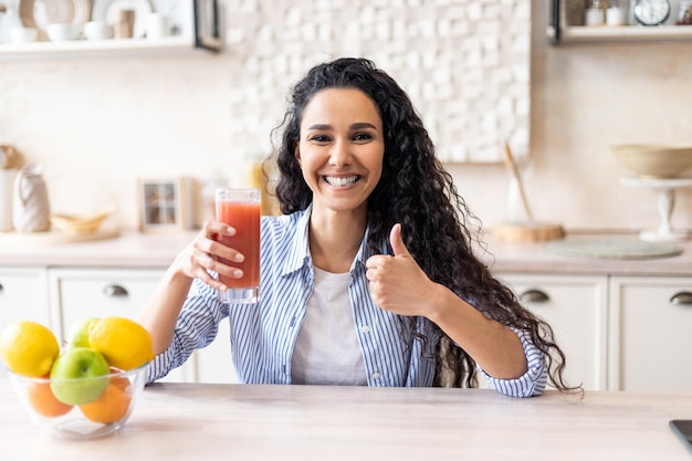 Femme latine positive tenant un verre de jus de tomate biologique et montrant le pouce assis à table dans