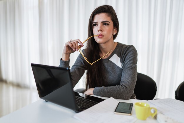 Femme latine réfléchie s'asseyant sur la table avec l'ordinateur portable pensant à l'inspiration rêvant