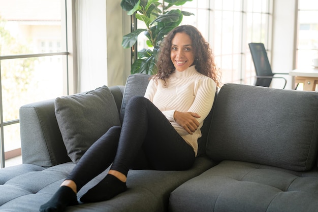 Femme latine souriante et assise sur un canapé à la maison