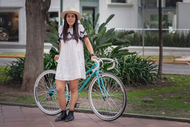 Femme latine avec un vélo portant une robe et un chapeau dans un parc