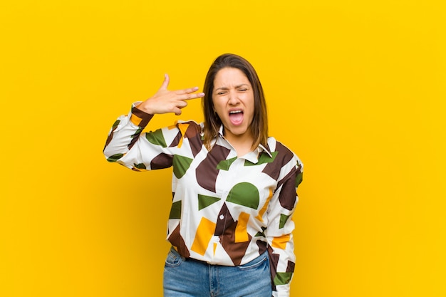 Femme latino-américaine à la malheureuse et stressée, geste de suicide faisant signe de pistolet avec la main, pointant vers la tête isolée contre le mur jaune