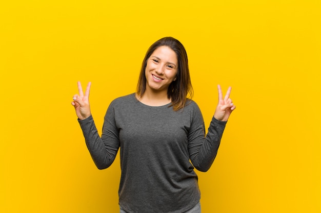 Femme latino-américaine souriante et semblant heureuse, amicale et satisfaite, victoire gesticulant ou paix à deux mains, isolée contre le mur jaune