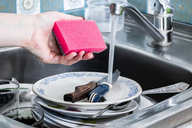 Une femme lave la vaisselle sale et tient dans sa main une éponge rose avec de la mousse.