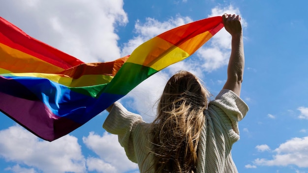 Femme lesbienne blonde tenant un drapeau d'identité de genre lgbt arc-en-ciel sur fond de ciel avec des nuages sur un