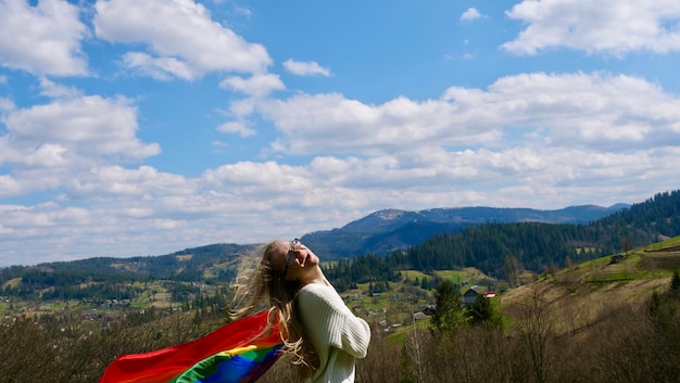 Femme lesbienne blonde tenant un drapeau d'identité de genre lgbt arc-en-ciel sur fond de ciel avec des nuages sur un