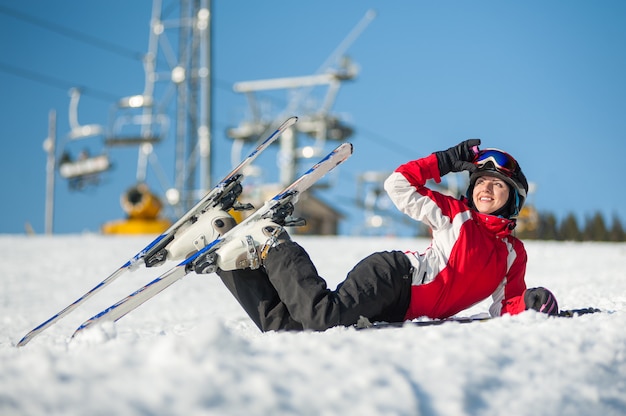 Femme levant ses lunettes de ski et regardant au loin, couchée avec des skis sur la neige au sommet de la montagne en journée ensoleillée