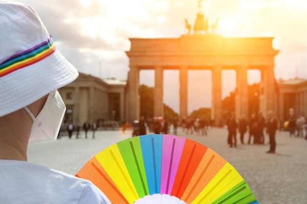 Femme LGBT caucasienne mature à Berlin avec ventilateur arc-en-ciel et ruban sur chapeau d'été blanc. Arc-en-ciel, symbole de la fierté gaie LGBTQIA, de la diversité. Femme portant un masque PP2. Ciel dramatique au-dessus de la porte de Brandebourg.