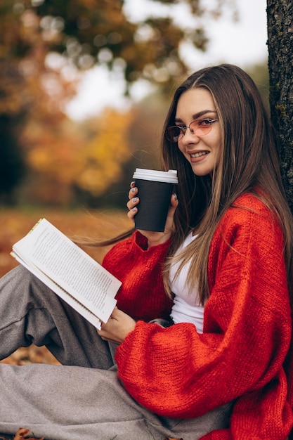 Femme lisant dans un parc d'automne et buvant du café sous l'arbre