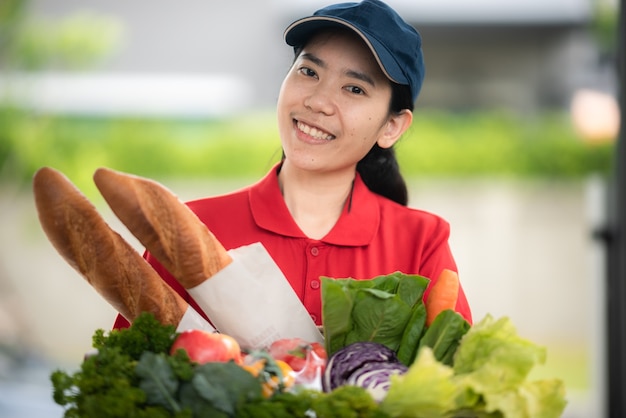 Femme de livraison asiatique en sac de manutention uniforme rouge de nourriture, fruits, légumes donnent au client devant la maison, concept de service de livraison