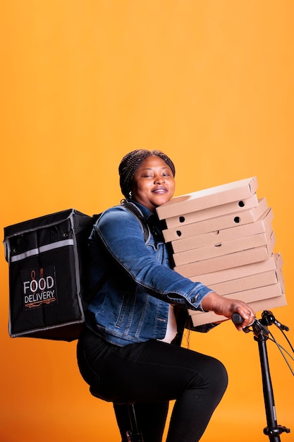 Femme de livraison à emporter souriante tenant une pile de boîtes en carton pleine de pizza livrant une commande de plats à emporter au client tout en faisant du vélo. Personne debout en studio sur fond jaune