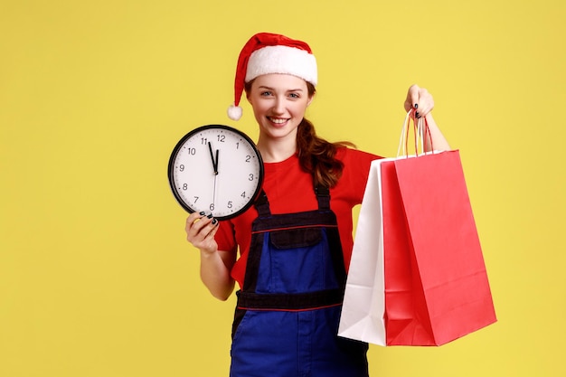 Femme de livraison heureuse souriante tenant une horloge murale et des sacs à provisions livraison rapide pour les vacances d'hiver portant une salopette bleue et un chapeau de père Noël Studio intérieur tourné isolé sur fond jaune