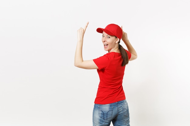 Femme de livraison en uniforme rouge isolé sur fond blanc. Jolie femme professionnelle en casquette, t-shirt, jeans travaillant comme coursier ou revendeur, pointant du doigt l'espace de copie pour la publicité. Vue arrière.