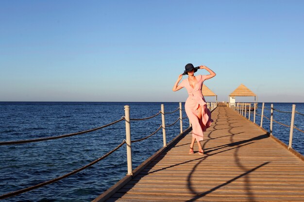 femme en longue robe rose marchant sur la plage dame en chapeau bleu paille et robe rose sur le port de bois