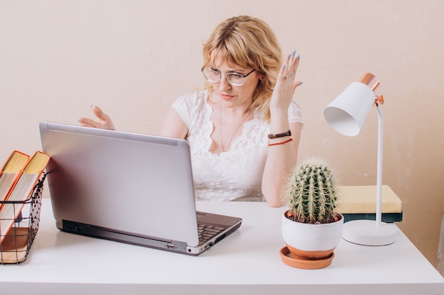 Une femme à lunettes et un chemisier blanc travaille sur un ordinateur portable et regarde avec surprise le moniteur