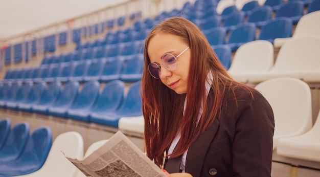Une femme à lunettes et un costume d'affaires lit un journal assis au stade