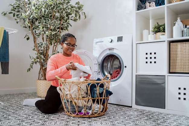 Une femme à lunettes effectue des tâches ménagères dans la buanderie de la salle de bain s'agenouille avec un panier en osier rempli de vêtements à la machine à laver charge des choses colorées dans le rinçage du tambour